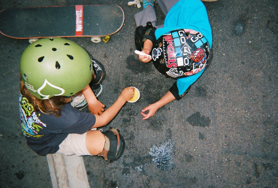 Overhead view of two kids drawing on the pavement in chalk beside a skateboard.